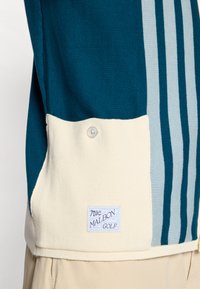 Teal strikket polo med kremfarget nederste del, prydet med tre lyseblå striper og en knappdetalj. Inkluderer en blå logopatch.