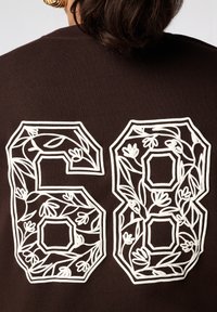 Bruine sweatshirt met grote witte cijfers "6" en "8" versierd met bloemmotieven. Het ontwerp bevat omtrekken met ingewikkelde plantmotieven.