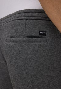 Nahaufnahme einer Person, die dunkelgraue Hosen mit einer Gesäßtasche und einem kleinen schwarzen Strellson-Label darüber trägt.