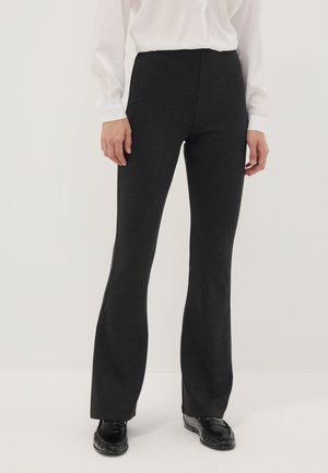 Persona que lleva pantalones negros acampanados ajustados, blusa blanca de manga larga y mocasines negros, de pie contra un fondo claro y liso.