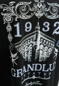 Sort t-shirt med hvid grafisk design af en bygning, blomsteraccenter, året "1932" og teksten "GRANDLUX ESTATES NEW YORK."