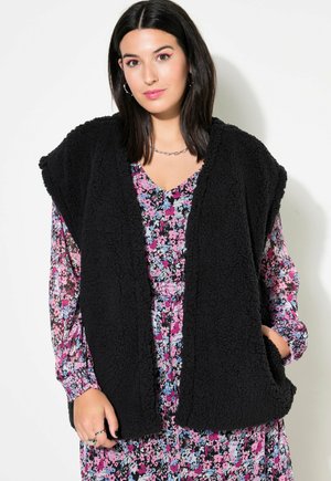 Schwarze Teddy-Stoff-Weste mit offenem Vorderteil, ausgestattet mit zwei seitlichen Taschen, getragen über einem blumigen Langarmkleid mit rosa, blauen und schwarzen Mustern.