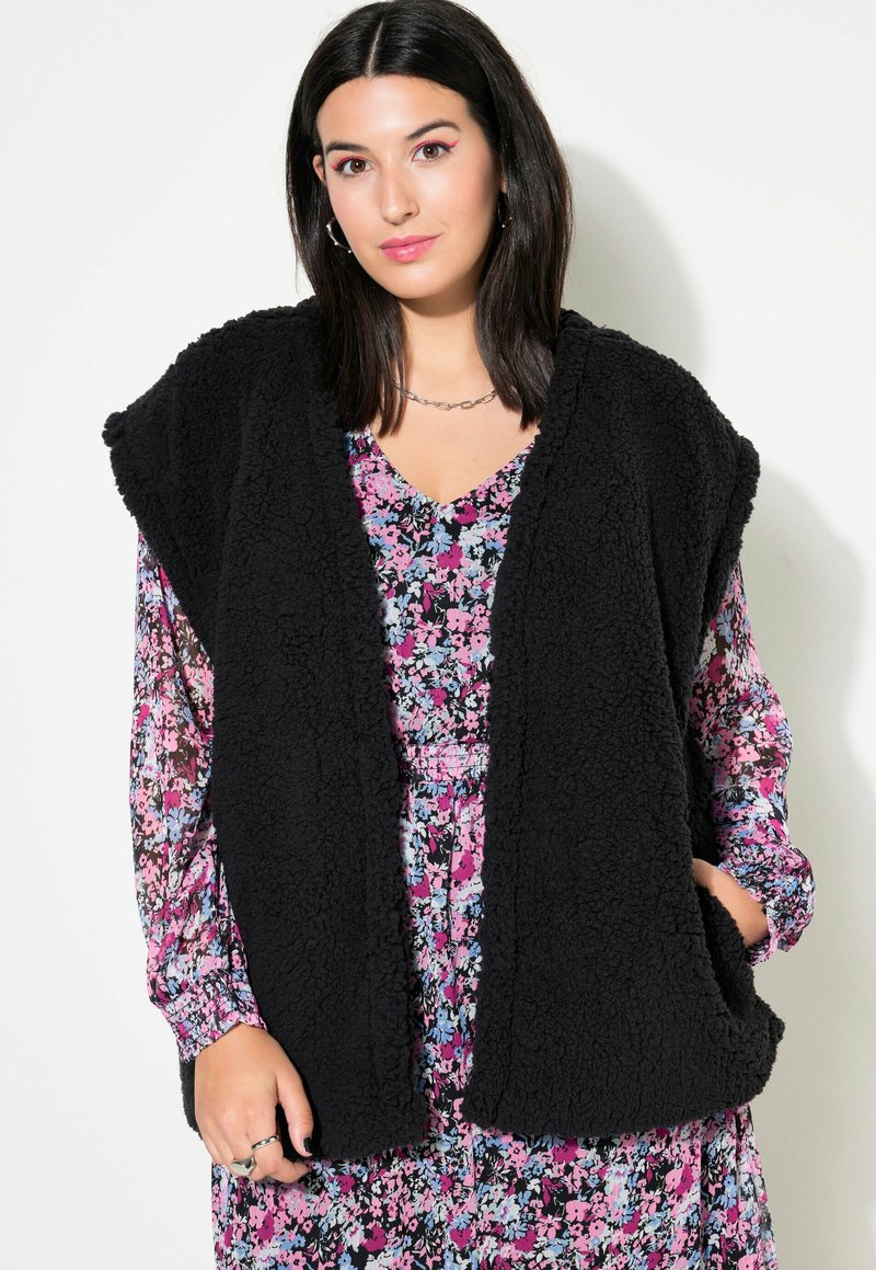 Schwarze Teddy-Stoff-Weste mit offenem Vorderteil, ausgestattet mit zwei seitlichen Taschen, getragen über einem blumigen Langarmkleid mit rosa, blauen und schwarzen Mustern.
