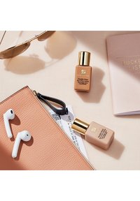 Zwei Estee Lauder Double Wear Foundationflaschen in Beige mit goldenen Kappen, neben weißen Ohrhörern, einem pinkfarbenen Texturbeutel, Sonnenbrillen und einem Notizbuch.