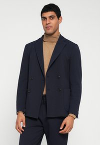 Ναυτικό διπλόσκαρδο σακάκι με μαύρα κουμπιά, υφή υφάσματος, πέτο με σχισμή και πλαϊνές τσέπες. Φορεμένο πάνω από ένα καμηλό ζιβάγκο.