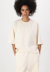 Beige oversized genser med raglanerm, med preget logo. Komplementert av matchende joggebukser med elastisk liv og aksentsting.