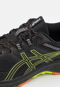Scarpa da ginnastica nera con accenti grigi riflettenti e logo lime vivace, caratterizzata da tessuto testurizzato e suola nera rinforzata.
