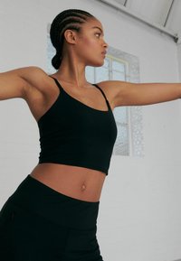 Top cropped negru, ajustat, cu bretele subțiri, asortat cu colanți negri cu talie înaltă, pe un fundal luminos și minimalist.