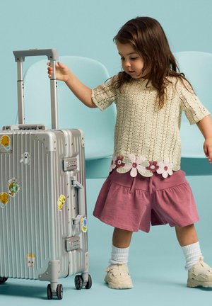 Jeune enfant en pull crème et short rose tire une valise argentée décorée d'autocollants colorés sur un fond bleu clair.