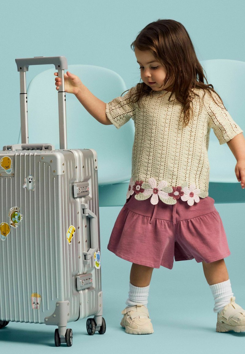 Jeune enfant en pull crème et short rose tire une valise argentée décorée d'autocollants colorés sur un fond bleu clair.