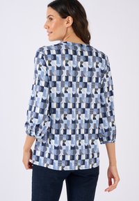 Blusa azzurra chiara con motivo geometrico e texture variate, con maniche a 3/4; abbinata a jeans blu scuro.