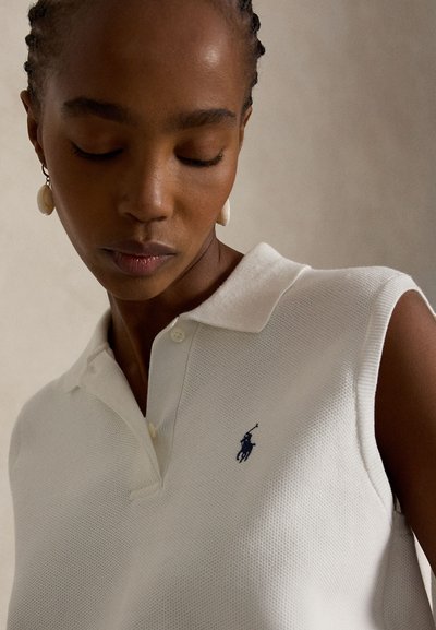 Moters su kasytėmis artimas vaizdas, kuri dėvi perlų auskarus ir baltą berankovį polo marškinėlį su mažu tamsiu siuvinėtu logotipu ant krūtinės.