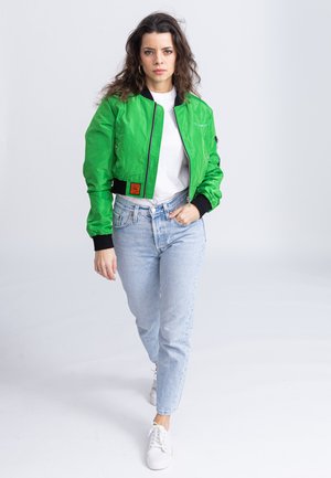 Groene bomberjack met een rits aan de voorkant, zwarte afwerking en twee zijzakken, gedragen met lichtblauwe spijkerbroek en witte sneakers.