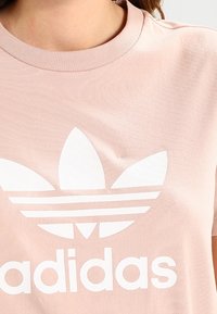 Ljusrosa bomullströja med rund halsringning, med en stor vit Adidas-logotyp och trefoil-design tryckt på framsidan.