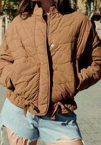 Personne portant une veste matelassée marron avec les mains dans les poches et un short en denim bleu clair déchiré à l'extérieur en plein jour.