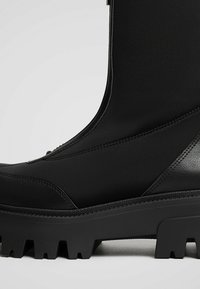 Botte noire imperméable dotée d'un matériau supérieur lisse, d'accents texturés et d'une semelle robuste avec des crampons prononcés pour une meilleure adhérence.