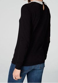 Svart långärmad tröja med en krage accentuerad av pärlor och en nyckelhål-design på baksidan. Kombinerad med blå denim jeans. Mjuk tygkvalitet.