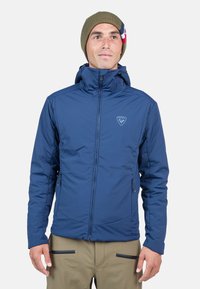 Marineblaue isolierte Jacke mit Kapuze, ausgestattet mit einem Frontreißverschluss, seitlichen Taschen und einem Logo auf der Brust. Glattes, wetterbeständiges Material.