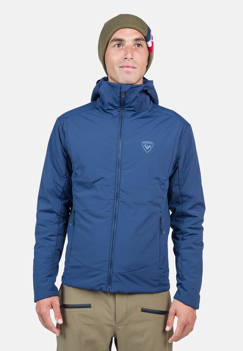 Marineblaue isolierte Jacke mit Kapuze, ausgestattet mit einem Frontreißverschluss, seitlichen Taschen und einem Logo auf der Brust. Glattes, wetterbeständiges Material.
