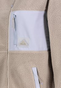 Beige fleece jacket with a light blue nylon pocket; features a subtle texture, side zip, and a triangular brand label in the pocket.