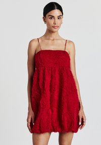 Frau in einem ärmellosen, roten strukturierten Minikleid mit dünnen Trägern und floralem Rosettenmuster, die vor einem schlichten hellen Hintergrund steht.