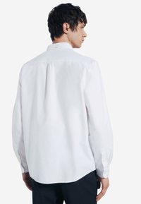 Camicia bianca a maniche lunghe con colletto a bottoni, in morbido cotone, con piega sul retro e polsini abbottonati. La camicia è indossata con pantaloni scuri.