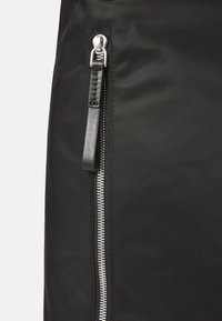 Close-up van een zwarte stoffen oppervlak met een verticale zilveren rits en een zwarte leren trekker.