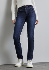 Mörkblå denimjeans med en figurnära design, som har fem fickor, synlig sömnad och en slät textur. Bärs med vita sneakers.