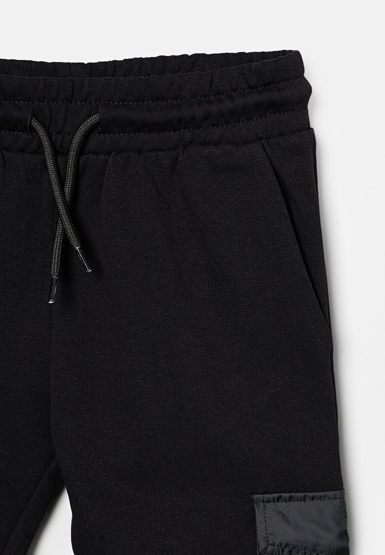 Pantalon de jogging noir avec taille élastique, accents de cordon de serrage, poches latérales et un patch en tissu contrasté sur la jambe droite.