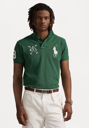 Groene poloshirt met een kraag met twee knopen, voorzien van een wit geborduurd logo "RPL" en gekruiste polo-stokken. G gedragen met witte broek en bruine riem.