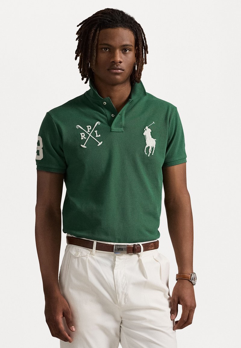 Grønn polo skjorte med to-knapps krage, med hvit brodert logo, "RPL," og kryssede polo staver. Båret med hvite bukser og brun belte.
