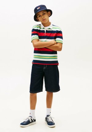 Jongen met een gestreepte polo, donkere spijkershort, witte sokken, sneakers en een marineblauwe vissershoed, staand met gekruiste armen tegen een witte achtergrond.