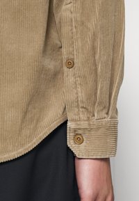 Camisa de pana beige con textura acanalada vertical, puños abotonados y dobladillo redondeado. Botones marrones que complementan el color de la tela.