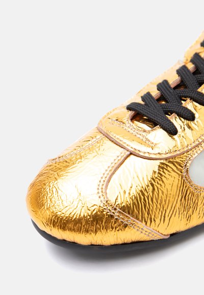 Λαμπερό χρυσό αθλητικό παπούτσι με ρυτιδωτή υφή, μαύρα κορδόνια, λεπτομερείς ραφές και μαύρη σόλα σε λευκό φόντο.