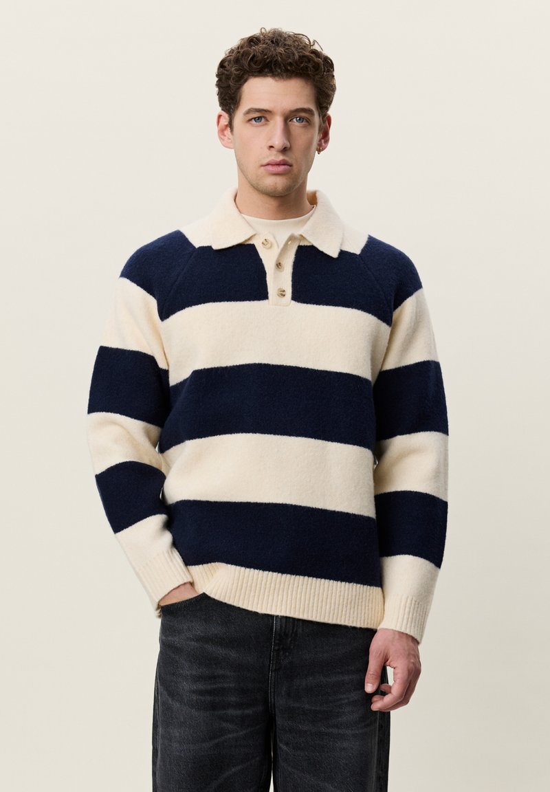 Jeune homme aux cheveux bruns bouclés, portant un pull à larges rayures crème et bleu marine, debout les mains partiellement dans les poches sur un fond uni.