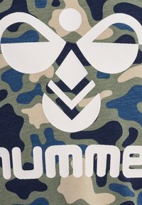 Tarnmusterstoff in Grün, Beige und Blau mit einem großen weißen Logo. Die Textur scheint gerippt zu sein und das Design umfasst geometrische Formen.