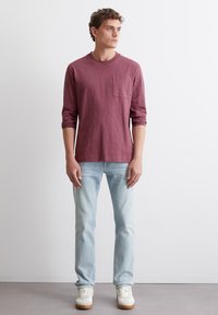 Langärmliges, kastanienbraunes T-Shirt mit Brusttasche, kombiniert mit hellblauen Jeans und weißen Sneakers, stehend auf einem neutralen Boden.