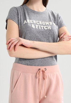 Osoba ubrana w szarą koszulkę Abercrombie & Fitch, z założonymi skrzyżowanymi ramionami, oraz różowe spodnie dresowe z sznurkiem na talii, na jednolitym tle.