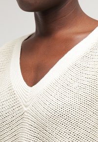 Pletený svetr s výstřihem do V v barvě off-white, zdobený texturovaným vzorem a žebrovanými detaily kolem výstřihu a lemu. Měkký, prodyšný materiál.