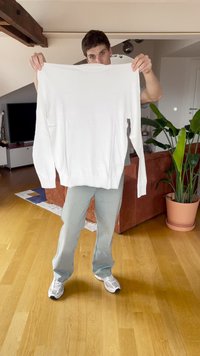 Maglione bianco a maniche lunghe sollevato, realizzato in un tessuto liscio. Presenta un colletto a girocollo e polsini a costine. Design leggermente strutturato e minimalista.