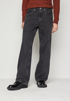 Schwarze, weite Jeans mit einem ausgewaschenen Muster, ausgestattet mit fünf Taschen, silberfarbener Hardware und einem Knopfverschluss. Kombiniert mit glänzenden schwarzen Schuhen.