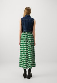 Veste sans manches en denim associée à une longue jupe verte avec un motif géométrique vert et blanc. Des bottes à plateforme noires complètent le look.