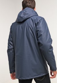 Marineblå vandtæt jakke med hætte, glat tekstur, lange ærmer og justerbare manchetknapper. Set bagfra med enkelt design.