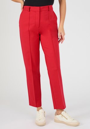 Pantalones rojos a medida de pierna recta combinados con zapatillas blancas bajas, de pie contra un fondo claro y liso.
