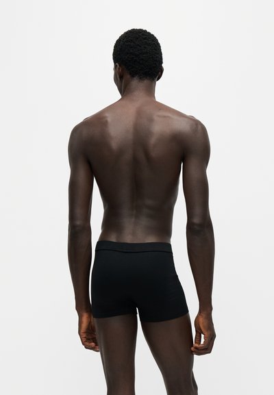 Homme à la peau foncée et aux cheveux courts bouclés, debout dos à la caméra, portant un short noir ajusté sur un fond clair uni.