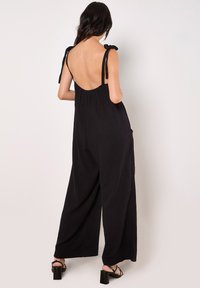 Black jumpsuit with wide legs, adjustable tie straps, and a deep back neckline. Made from lightweight fabric with a textured finish.