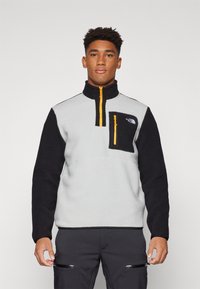 Fleece pullover in lichtgrijs en zwart, met een kwart rits, hoge kraag en een met rits afsluitbare borstzak met felle oranje details.