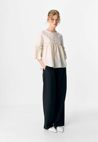 Blouse beige clair à manches courtes volantées et ourlet froncé, associée à un pantalon noir ample et des chaussures blanches pointues.