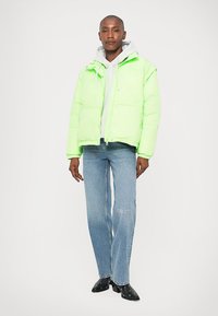 Νεον πράσινο μπουφάν με φερμουάρ, συνδυασμένο με ελαφριά μπλε τζιν παντελόνι με λευκή κεντητή επιγραφή. Μαύρες γόβες ολοκληρώνουν την εμφάνιση.