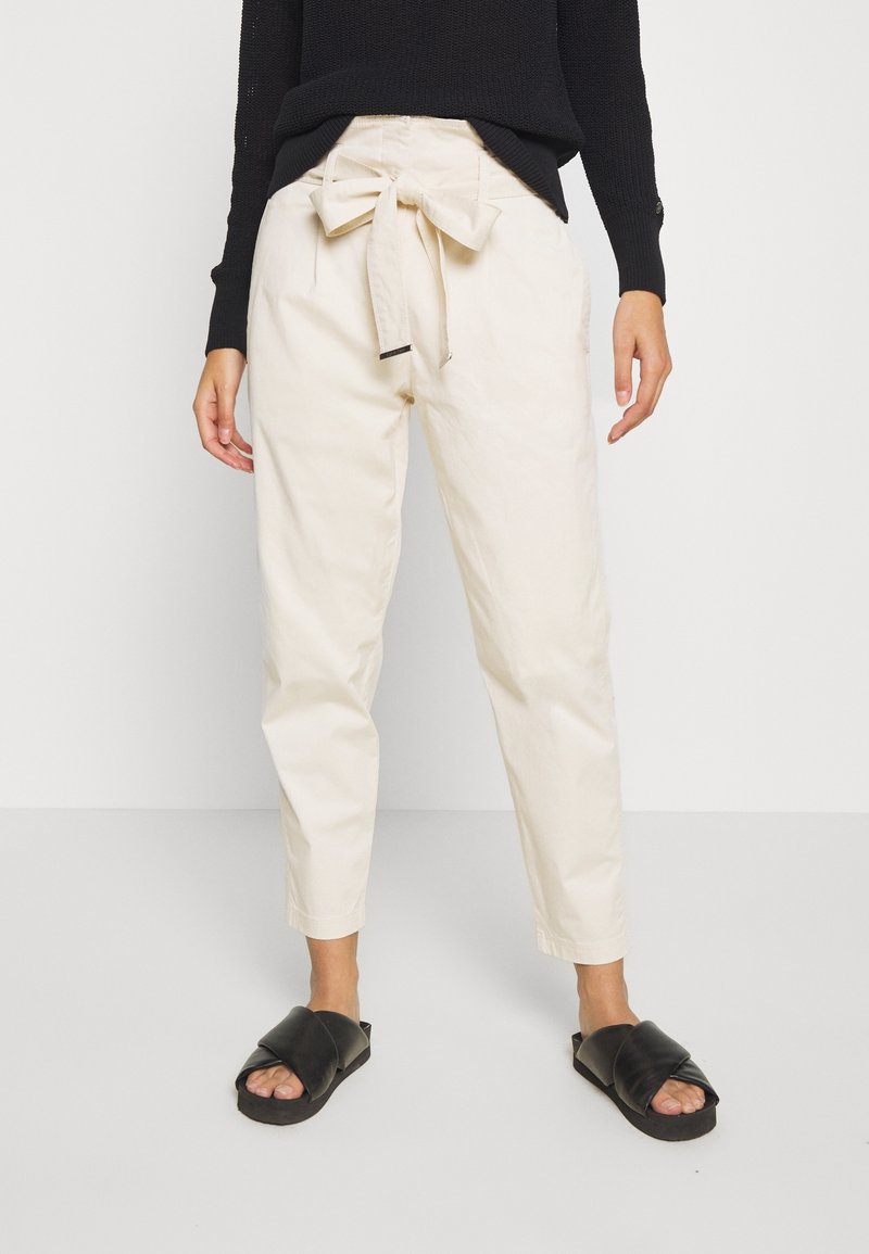 Personne portant un pantalon beige taille haute avec un nœud devant, un haut noir à manches longues et des sandales noires à bouts ouverts, debout sur un sol uni.